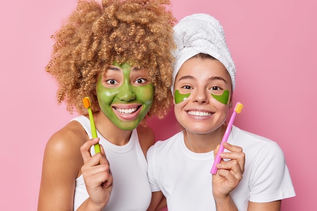 Indoor shot of positive young women apply green beauty mask and hydrogel patches hold toothbushes take care of teeth dressed in casual clothes isolated over pink background Oral hygiene concept