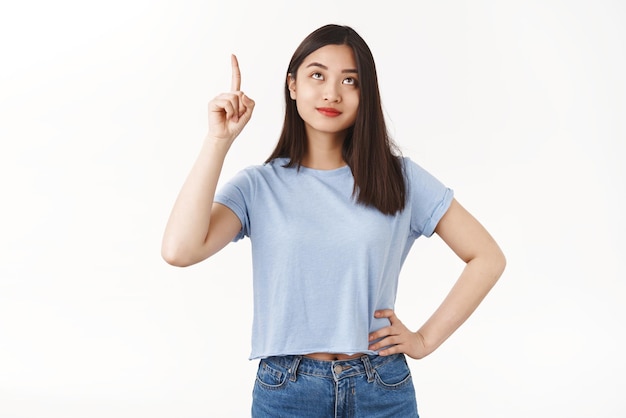 Indoor shot pretty asian brunette female look up pointing top smirking see sibling come from upstairs standing white background curious contemplating awesome summer promo offer