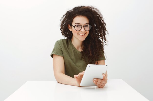 Indoor shot of pretty brunette in black glasses, sitting at white table and browsing in network via digital tablet