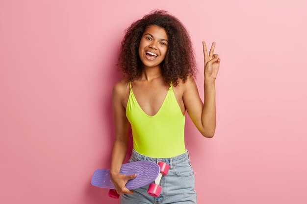 Indoor shot of pretty curly haired woman with glad expression, raises hand and makes peace sign, wears yellow vest and denim shorts, has slim figure, holds skateboard, enjoys favourite hobby