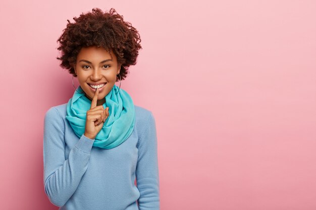 Indoor shot of pretty dark skinned woman asks keep quiet, makes shush gesture, wears blue sweater and scarf over rosy wall
