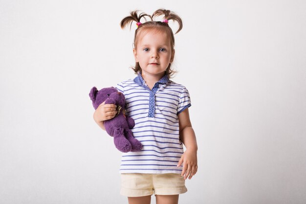 Indoor shot of pretty little child in t shirt and shorts, holds purple teddy bear in hands