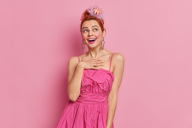 Indoor shot of pretty redhead young woman smiles gladfully keeps hand on chest