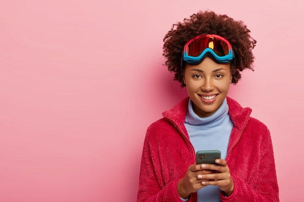 Indoor shot of pretty skier woman uses smartphone device, wears ski mask, smiles happily at camera, isolated over pink studio wall.