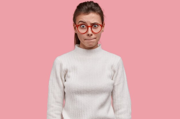 Indoor shot of pretty woman presses lips, raises eyebrows in bewilderment, reacts on something unexpected, wears white sweater