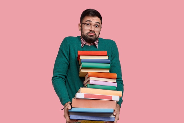 Indoor shot of puzzled unshaven man wears spectacles, holds many handbooks, feels doubt