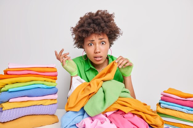 어리둥절한 젊은 아프리카계 미국인 여성의 실내 사진은 세탁을 위해 옷을 고르고 집안일을 하느라 바쁜 흰색 벽에 기대어 테이블에 앉아 깔끔한 옷 더미를 만듭니다.