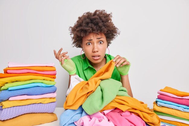 Indoor shot of puzzled young African American woman picks clothes for washing folds laundry busy doing housekeeping chores sits at table against white wall makes neat piles of clothing