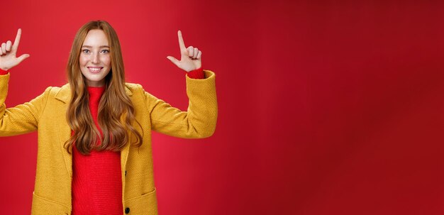 Снимок в помещении рыжеволосой привлекательной женщины в желтом осеннем пальто, поднимающей руки, продвигающей рекламу