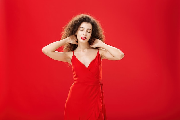 Free photo indoor shot of relaxed woman messaging back of neck with closed eyes and calm carefree expression standing in elegant evening dress with curly hairstyle against red background pleased and tired.