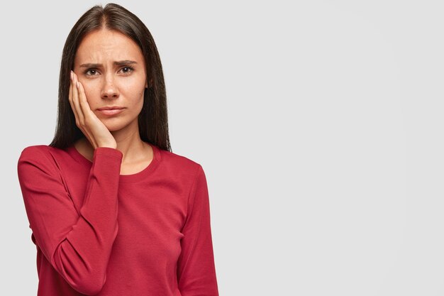 Indoor shot of sad unhappy European woman with unhappy expression, keeps palm on cheek, wears casual red sweatshirt,