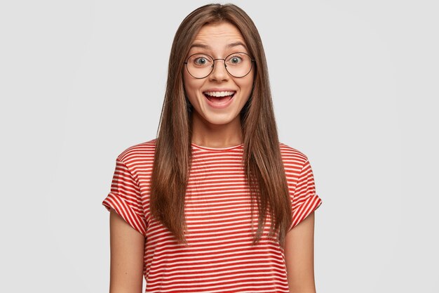 Indoor shot of satisfied brunette woman with dark hair, wears round spectacles, smiles joyfully