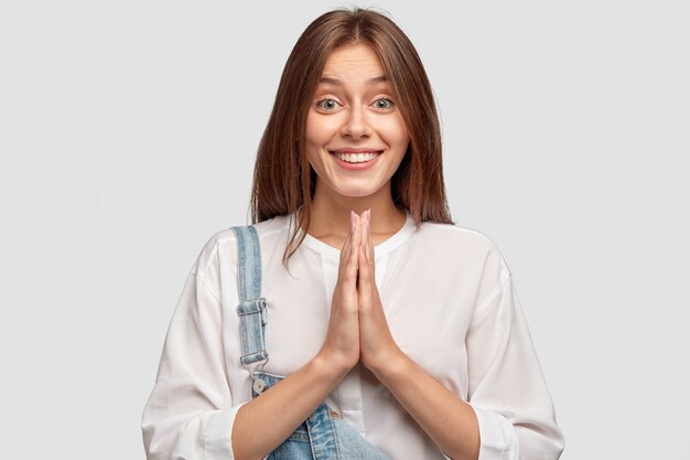 Indoor shot of satisfied good looking woman begs for mercy, has positive expression, gentle smile, keeps palms in praying gesture