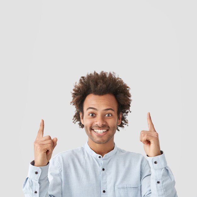 Indoor shot of satisfied happy African American male with curly hair, has broad shining smile, points upwards with both index fingers