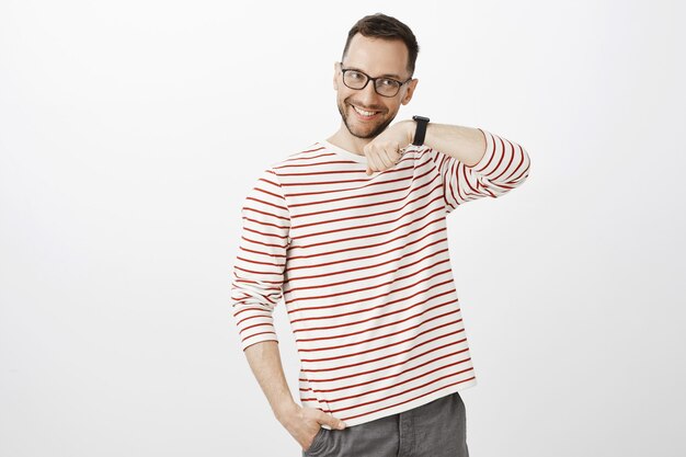 Indoor shot of satisfied happy european man with bristle in glasses, raising hand and listening to watch sound, being pleased with quality of gadget