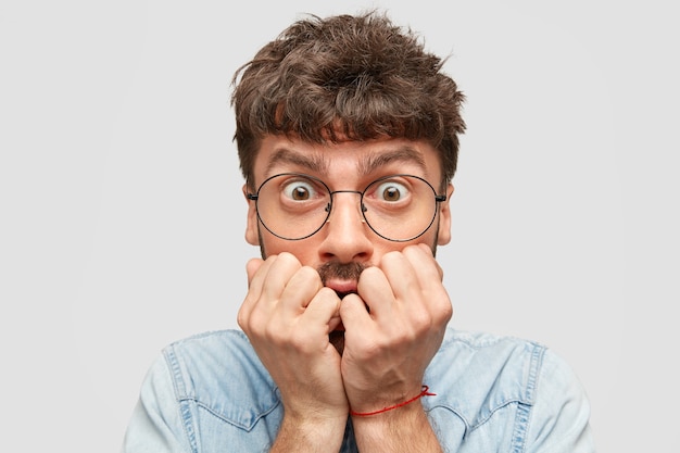 Indoor shot of scared nervous man student bites finger nails, stares with fear, has dark hair