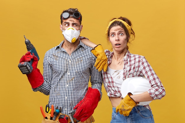 Free photo indoor shot of scared woman wearing protective gloves holding hardhat leaning at her husband's shoulder who is going to fix shelves in room. untidy handyman and his cute wife