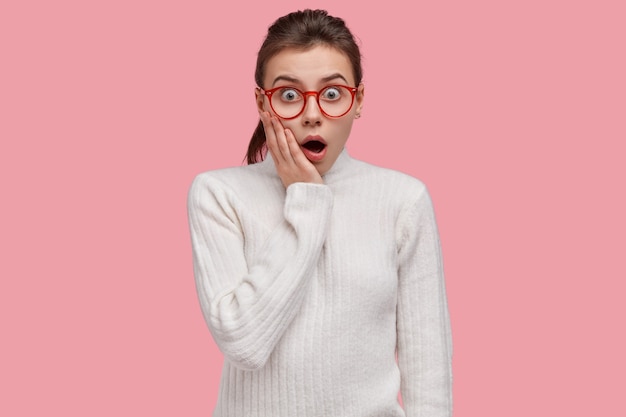 Free photo indoor shot of scared young european woman keeps jaw dropped, touches cheeks, wears casual white jumper