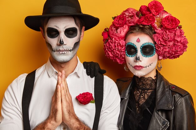頭蓋骨の化粧をした怖いカップルの屋内ショット、伝統的なメキシコの服を着て、死者の日カーニバルを訪れ、不気味な顔をして、男性は祈りのポーズで立っています