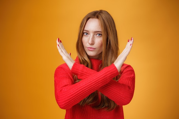 Снимок уверенной в себе привлекательной мощной женщины с рыжими волосами и веснушками, скрестив руки в помещении, запрещающий жест, запрещающий совершать действия, уверенно стоя над оранжевой стеной
