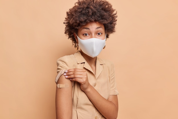 Indoor shot of serious Afro American woman with curly hair got her vaccination dose protects against coronavirus shows vaccinated arm with adhesive plaster disposable face mask isolated on beige wall