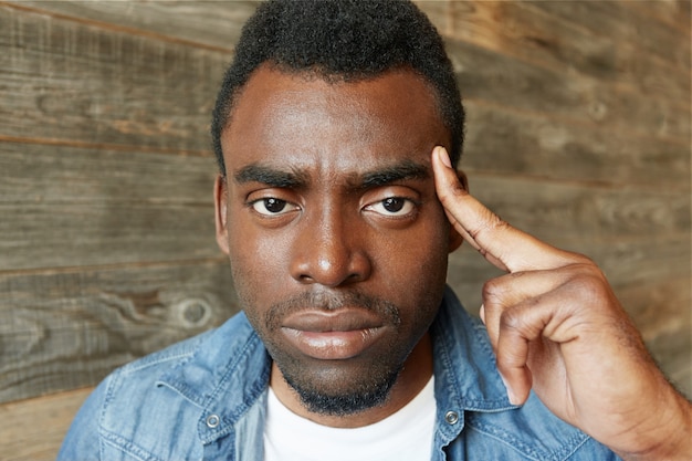 Free photo indoor shot of serious fed-up bearded african male in jeans jacket holding finger on his temple, frowning and having angry face as if saying: use your brain, stop talking nonsense. body language