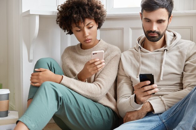 Indoor shot of serious man and woman use mobile phones for surfing internet, make shopping online