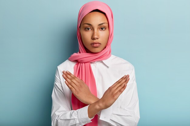 Indoor shot of serious Muslim woman makes denial hand gesture, keeps arms crossed over chest, demonstrates stop sign, wears headscarf, follows religious dress code, isolated over blue wall