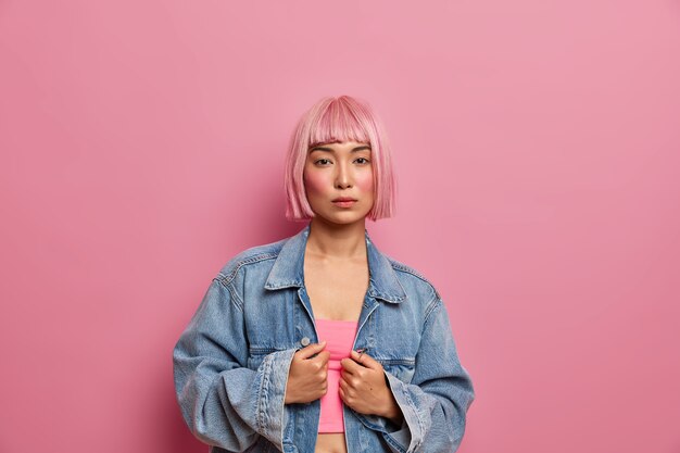 Indoor shot of serious pink haired female model dressed in top and oversized denim jacket, looks directly, wears minimal makeup, 