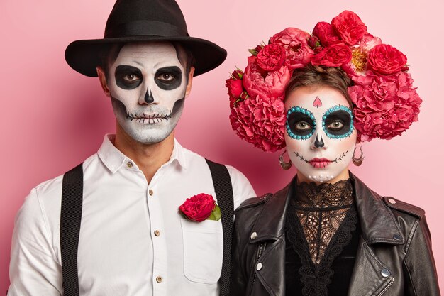 Снимок в помещении серьезной романтической пары, позирующей перед праздником Хэллоуина, в цветочном венке и шляпе на головах, в традиционных страшных костюмах, смотрящей прямо в камеру, с макияжем зомби в мексиканском стиле