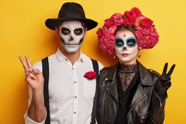 Indoor shot of serious woman and man in special scary costumes, make peace victory gesture, have vivid makeup for looking horrible, celebrate traditional holiday in Mexico