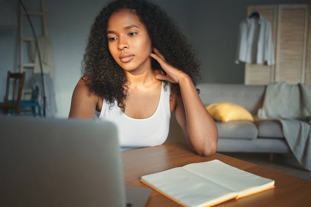 真面目な若いアフリカ系アメリカ人女性ジャーナリストがホームオフィスで働いて、ポータブルコンピューターを使用して、空白のコピースペースページのある開いた日記で机に座っている白いタンクトップの屋内ショット