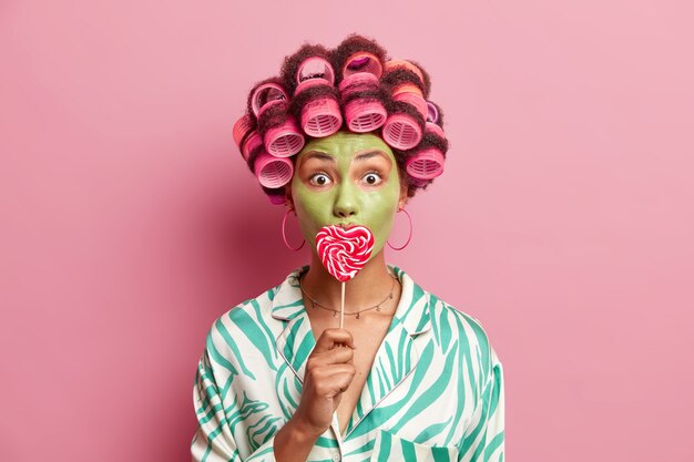 Indoor shot of shocked African American woman applies hair rollers to make perfect hairstyle covers mouth with lollipop makes green nourishing mask to reduce fine lines isolated over pink wall.