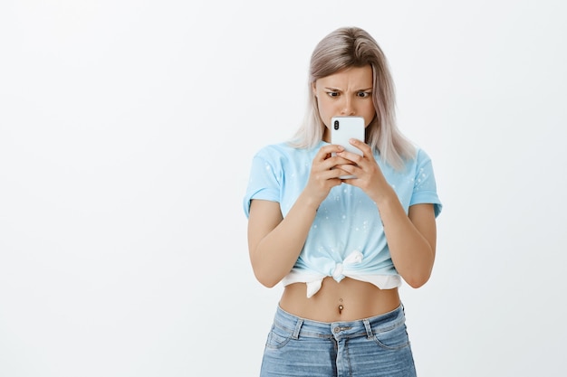 Indoor shot of shocked blonde girl posing in the studio with her phone