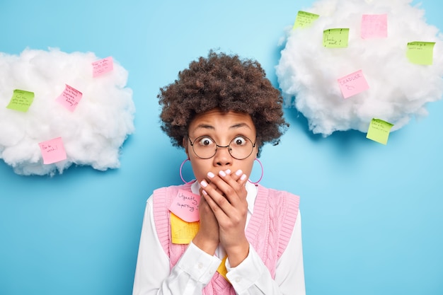 Free photo indoor shot of shocked ethnic female office worker keeps hands on mouth writes tasks and creative ideas on sticky post it notes realises deadline wears round spectacles