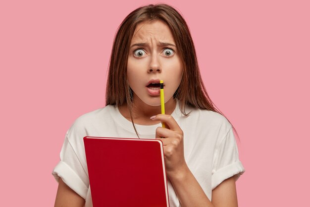 Indoor shot of shocked European woman stares with surprisement at camera, keeps mouth opened, holds pencil near mouth