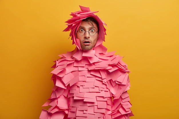 Indoor shot of shocked young Caucasian man stares with bugged eyes, wears transparent glasses, has creative costume made of sticky notes, stares startled and impressed, isolated over yellow wall