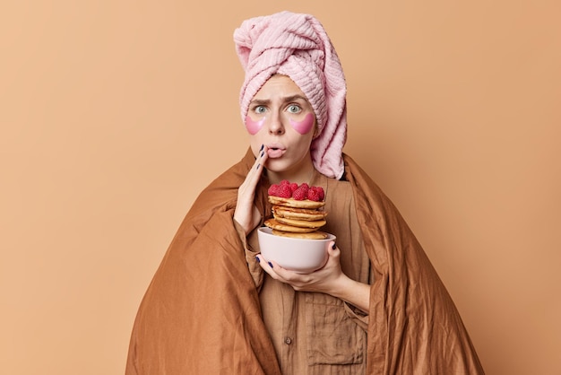 Indoor shot of shocked young woman looks wondered at camera wrapped in duvet holds bowl of pancakes hears shocking news in morning isolated over brown background applies beauty patches under eyes