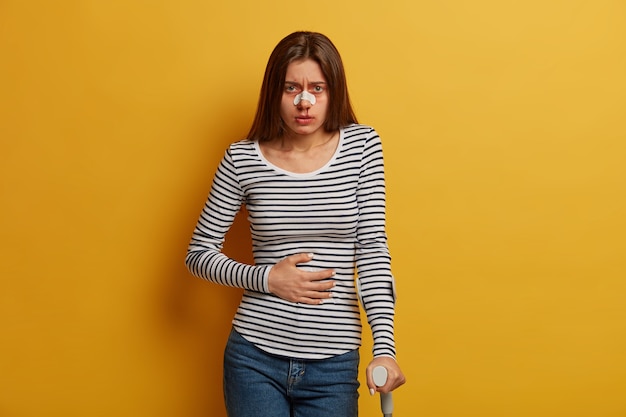 Foto gratuita ripresa in interni di una donna malata vittima di guida spericolata avverte fastidio allo stomaco, naso rotto e gamba, usa ausili per la mobilità, guarda tristemente, viene dal chirurgo, si riprende dopo il disastro