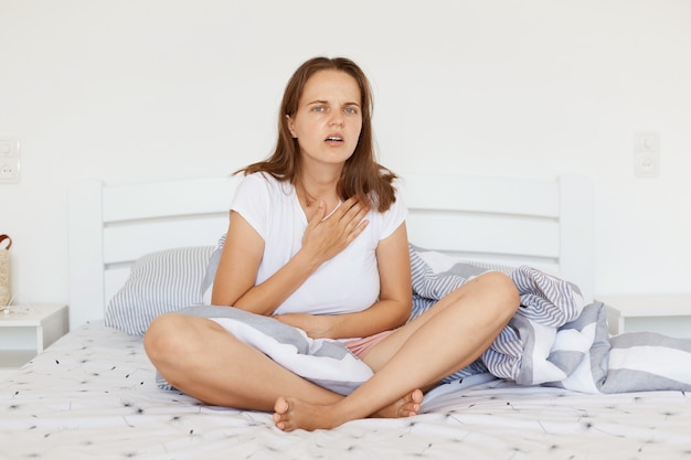 Indoor shot of sick woman wearing white casual t shirt, sitting on bed with crossed legs, touching her chest, suffering from heart pain, looking at camera with frowning face.