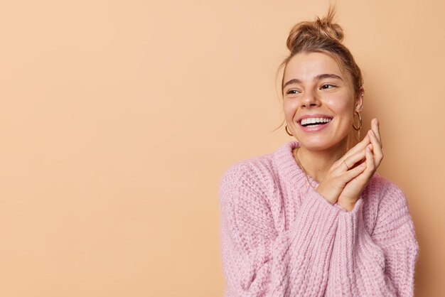 Indoor shot of sincere European woman with hair bun smiles broadly keeps hands pressed together feel very glad being in good mood focused away isolated over brown background copy space for your text