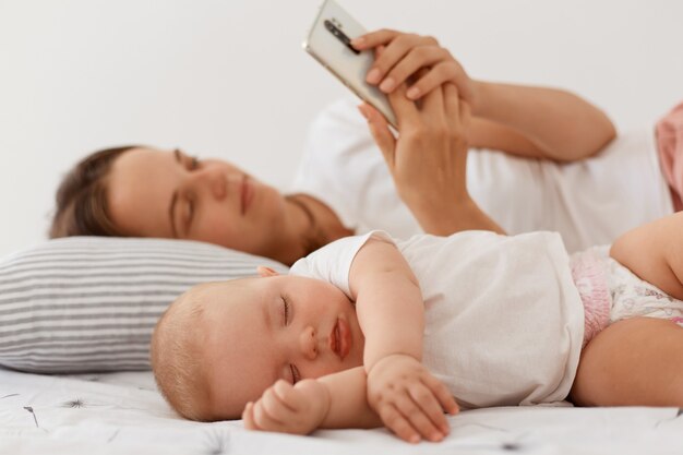 Indoor shot of sleeping baby wearing white t shirt, mother lying near daughter and holding cell phone, woman using smart phone for browsing internet or typing messages.