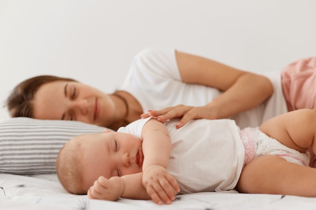 Indoor shot of sleeping woman and her charming small daughter lying on bed with closed eyes, resting in afternoon, mommy looking at baby with great love and hugging her.