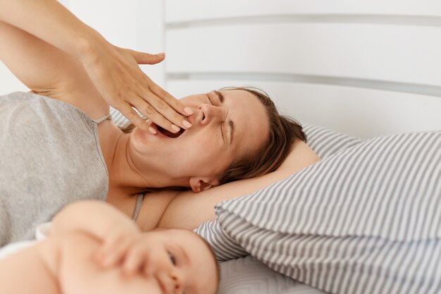 Indoor shot of sleepy mother lying in bed with infant baby daughter or son, woman yawning, covering mouth with hand, has sleepless night, needs energy.
