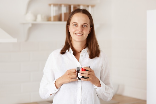웃고 있는 아름다운 여성이 부엌에서 커피 한 잔을 마시고 행복한 표정으로 카메라를 바라보는 실내 사진, 흰색 캐주얼 스타일 셔츠를 입은 여성.