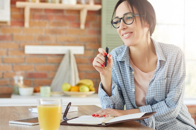 Indoor shot of smiling happy woman sits at kitchen table, makes notes in diary, plans what to do,