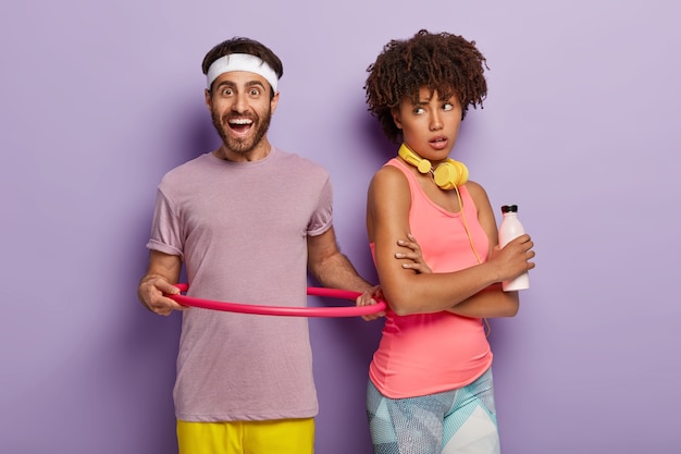 Indoor shot of smiling man rotates hula hoop, dressed in purple t shirt, being in good physical shape, Afro woman stands back, holds bottle of fresh water, isolated over purple wall. Healthy lifestyle