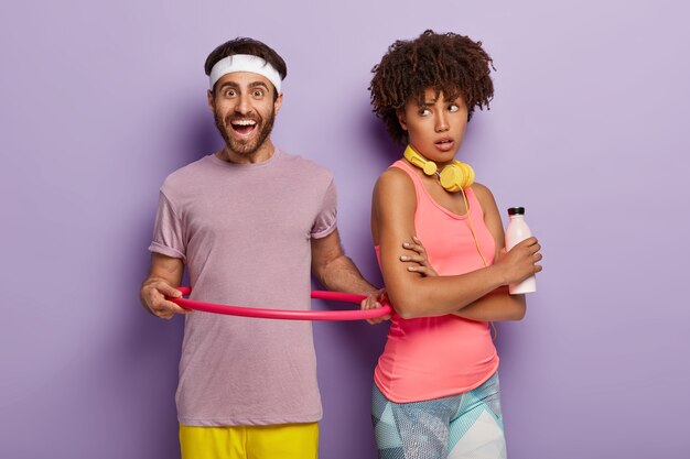 Снимок в помещении: улыбающийся мужчина вращает обруч, одетый в фиолетовую футболку, находится в хорошей физической форме, афро-женщина стоит в стороне, держит бутылку пресной воды, изолированную над фиолетовой стеной. Здоровый образ жизни