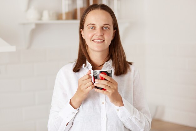 Крытый снимок улыбающейся красивой женщины с чашкой кофе на кухне, стоящей с приятной улыбкой, наслаждающейся горячим чаем утром после завтрака, смотрящей в камеру с позитивом.