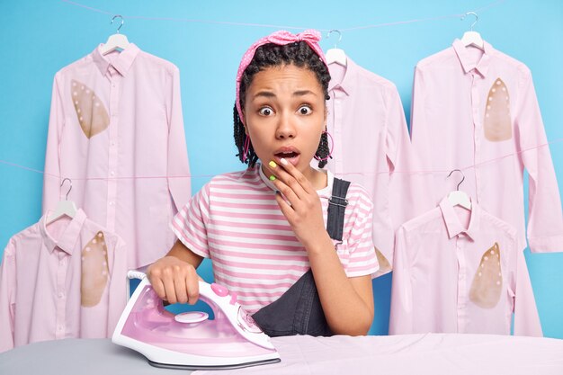 Indoor shot of startled young Afro American housewife stares surprised at camera holds breath from wonder stands near ironing board irons clothes poses in laundry room busy doing domestic work